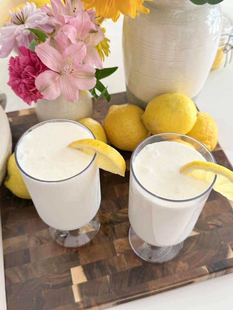 Two glasses of whipped frozen lemonade garnished with lemon slices, surrounded by fresh lemons and pink flowers on a wooden tray.