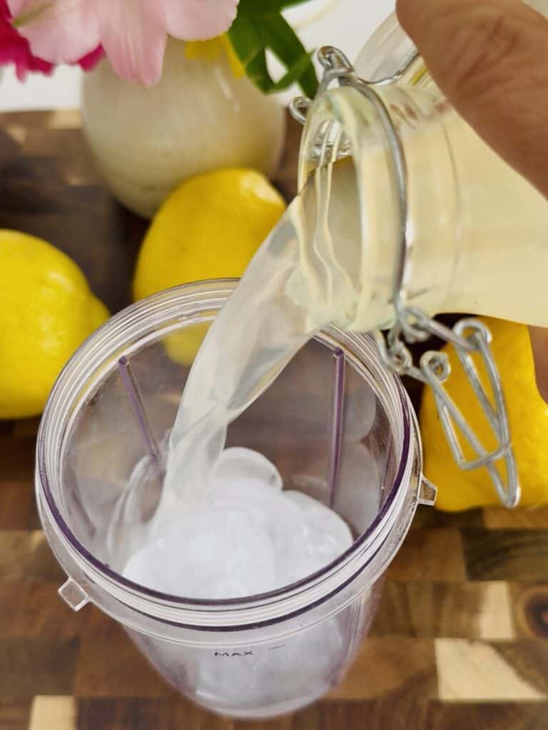 Adding lemonade to a clear container containing ice around by lemons and pink flowers, all in a dark brown cutting board