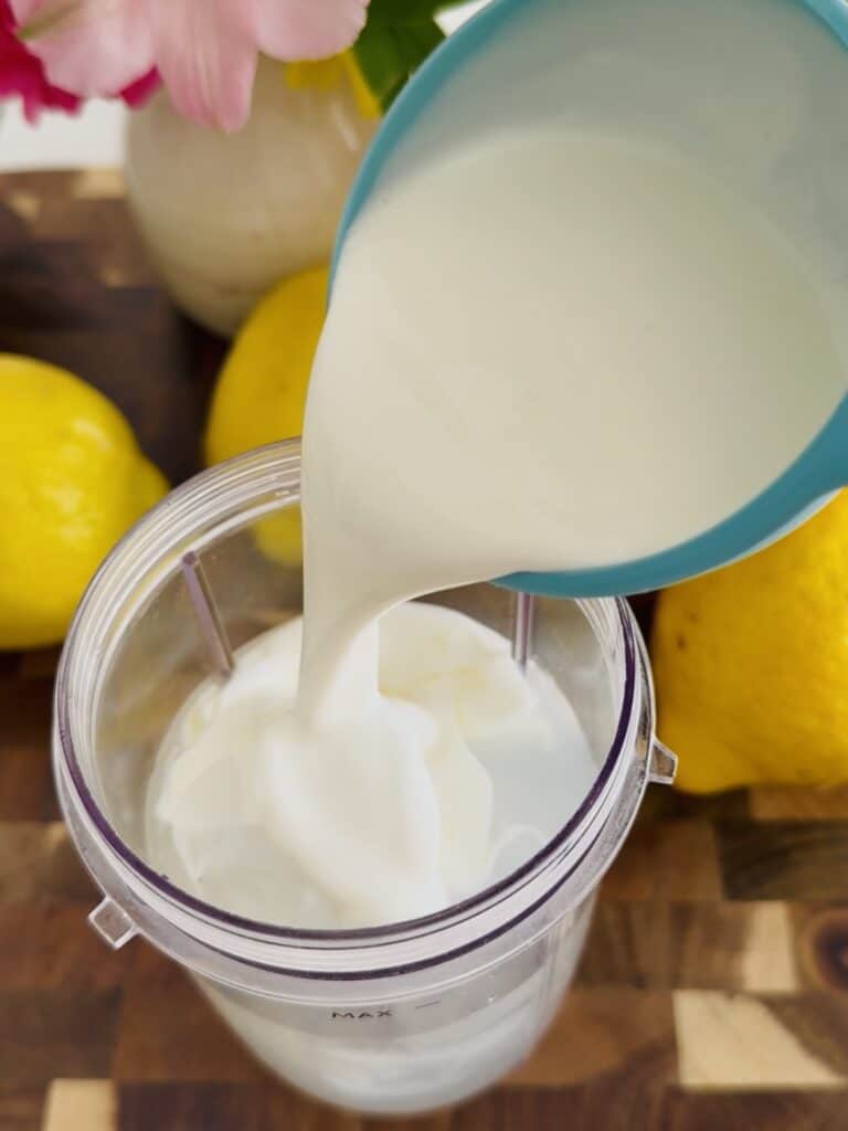Adding heavy whipping cream that is in a blue one cup measuring cup into a clear container that is containing lemonade, surrounded by lemons and pink flowers all in a dark brown cutting board