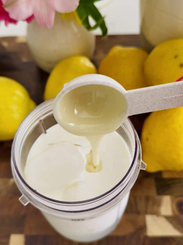 With a measuring spoon, add 1 tablespoon, of sweetened condensed milk into a container with lemonade and whip whipping cream that is in a clear container on a brown cutting board, surrounded by lemons and pink flowers