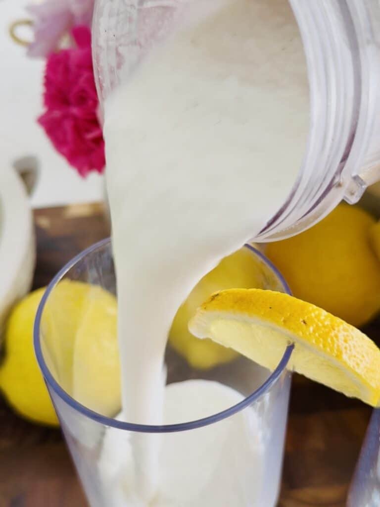 Pouring whipped frozen lemonade into a plastic clear glass, garnished with a sliced lemon on a brown cutting board for buy lemons and pink flowers