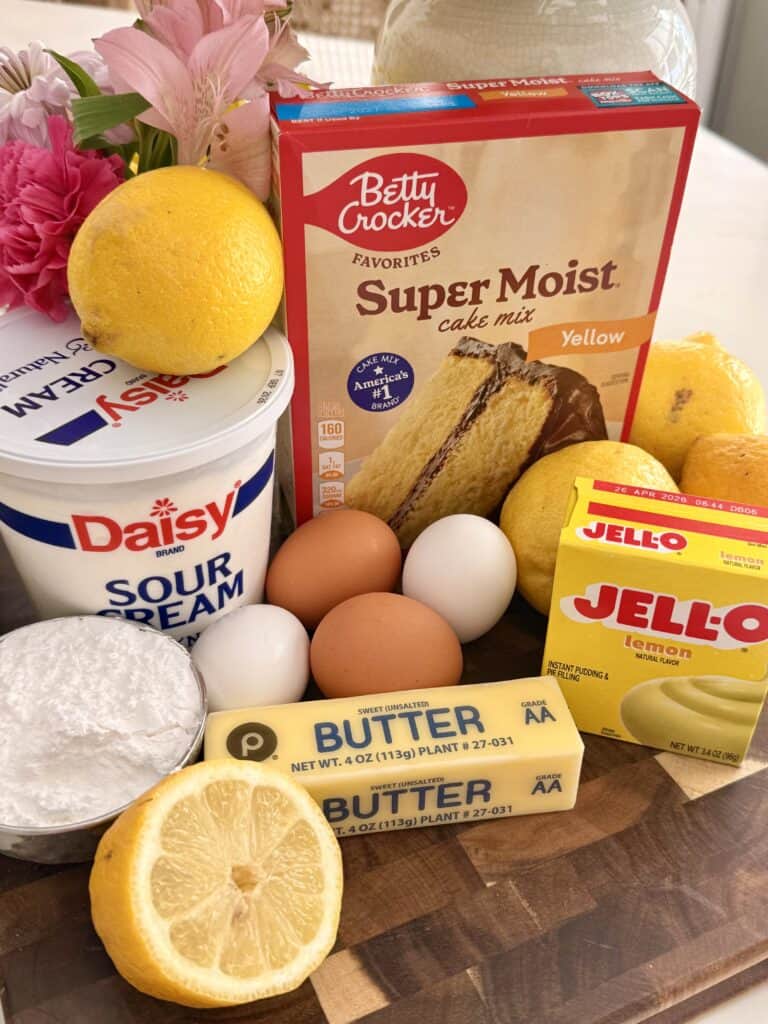 The ingredients in a duck brown cutting board they're super moist, Betty Crocker, yellow cake, lemons Jell-O, lemon pudding, four eggs to a brown to white unsalted butter, half a lemon, powdered sugar in a silver bowl on the counter and in the background is pink flowers in a vase
