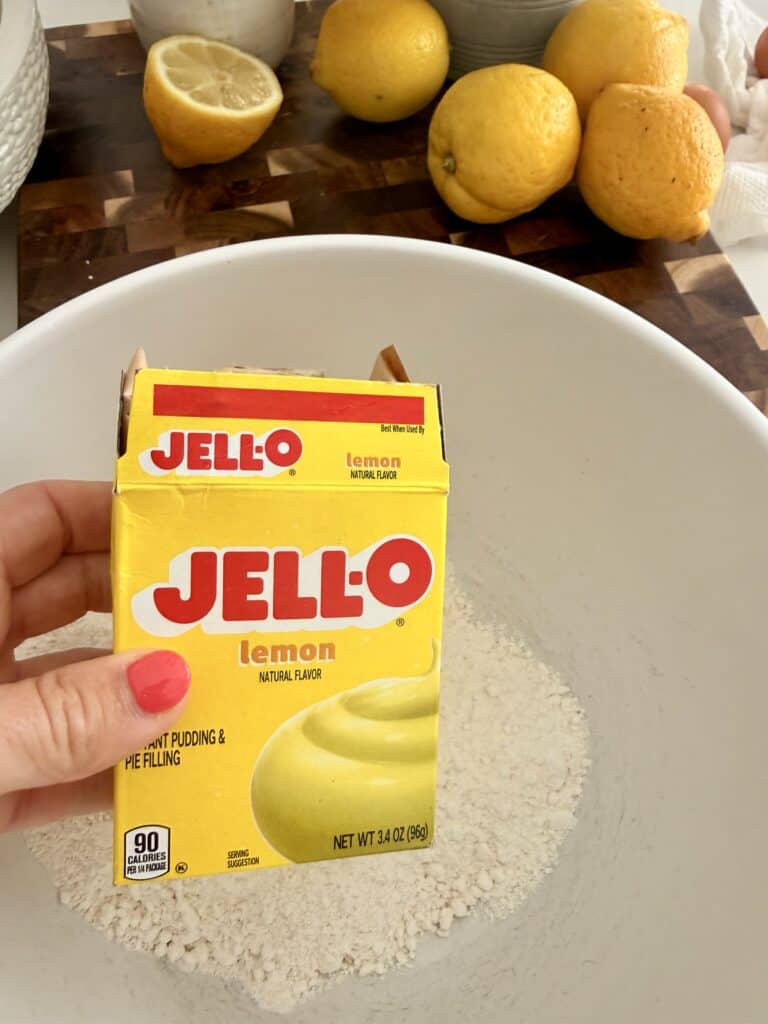 Holding a yellow Jell-O lemon instant pudding mix above a white mixing bowl with cake, yellow cake mix in it thrown by lemons and a dark brown cutting board