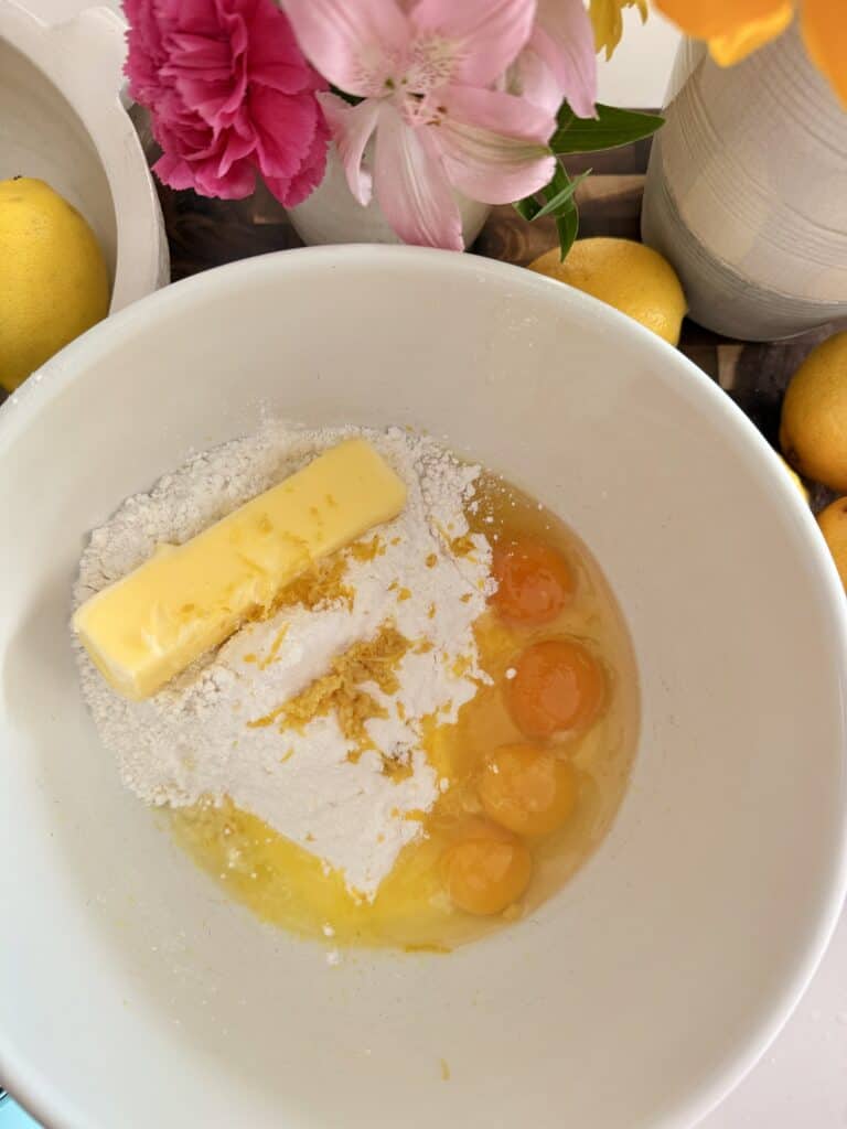 In a large white mixing bowl are four eggs, lemon juice, lemon sauce, cake mix Jell-O mix, sour cream and unsalted softened butter, all in a large white mixing bowl on the counter with surrounded by lemons, dark brown, cutting board and yellow and pink flowers