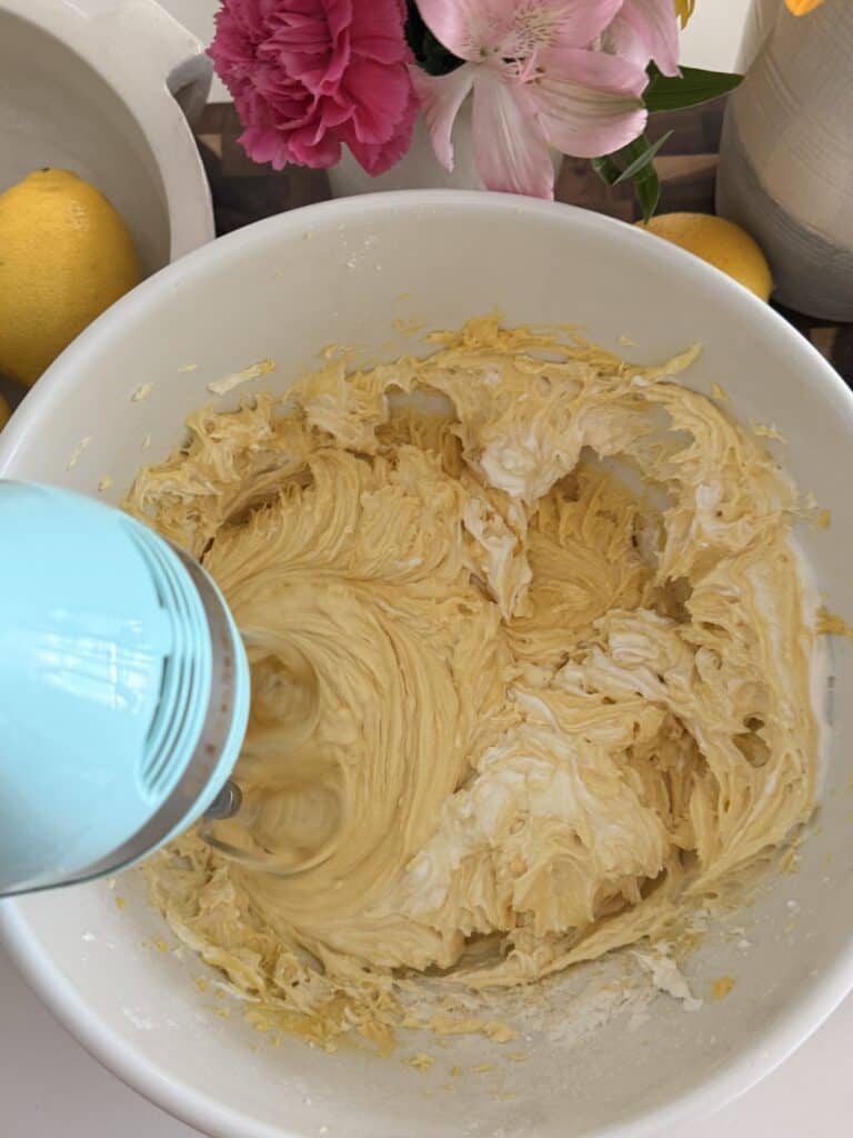 Using an electric mixer that is blue mixing all the cake ingredients in large white bowls round by a bowl of lemons and pink flowers
