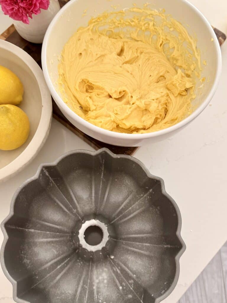 The mixed lemon pound cake cake mix is in a large white bowl and in front of that is the prepared and greased Bundt pan on the kitchen counter in front of Bible of lemons and pink flowers.