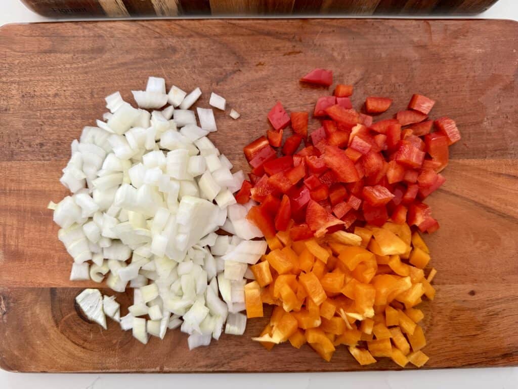 Chopped white onions, red bell peppers, and orange bell peppers on a wooden cutting board for Chicken Fajita Penne Pasta.