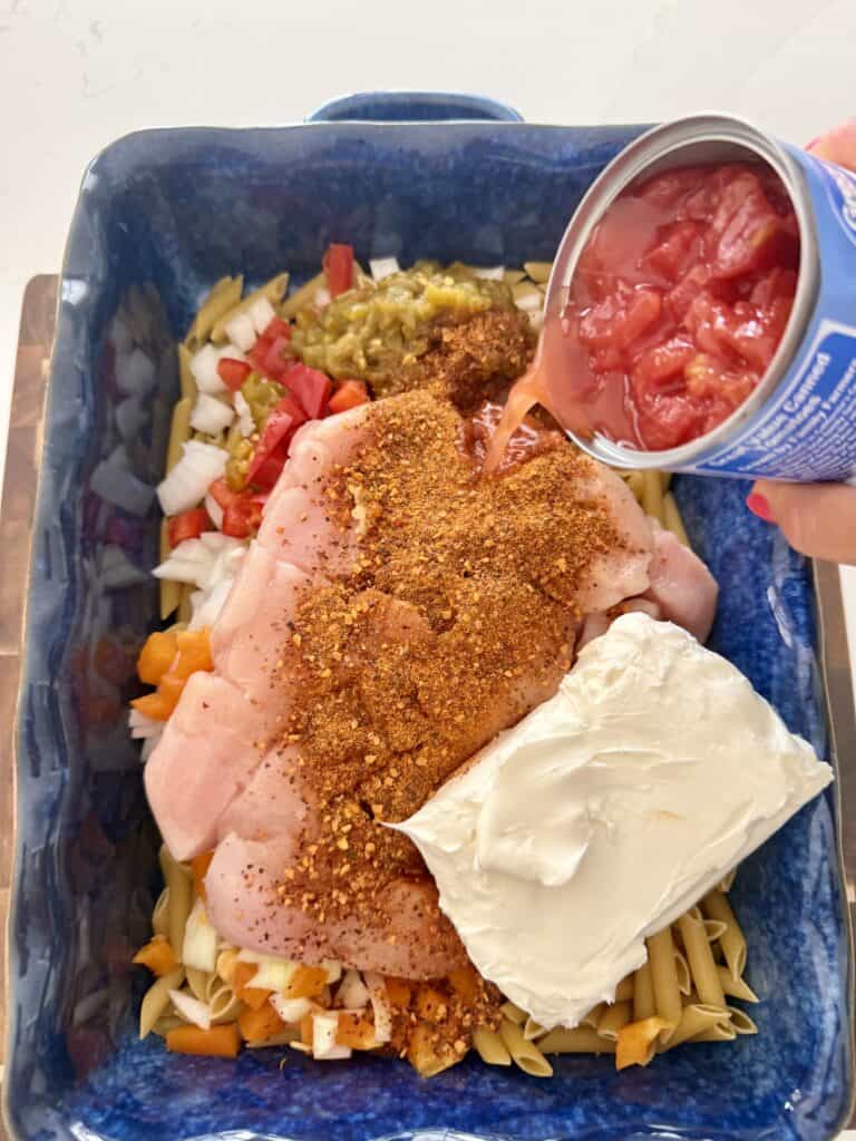Pouring a can of diced tomatoes over top of fajita seasoning, block of cream cheese, diced chicken, veggies, and pasta, and a blue casserole dish on the counter