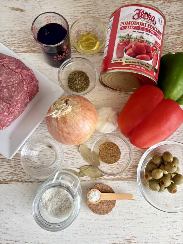 Assorted fresh ingredients for Picadillo including canned tomatoes, red and green peppers, onion, garlic, ground beef, olives, and spices.