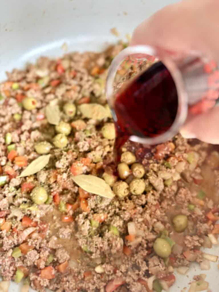 Person pouring red wine into a skillet with ground meat, tomateos, peppers, onions and spices for a flavorful dish.