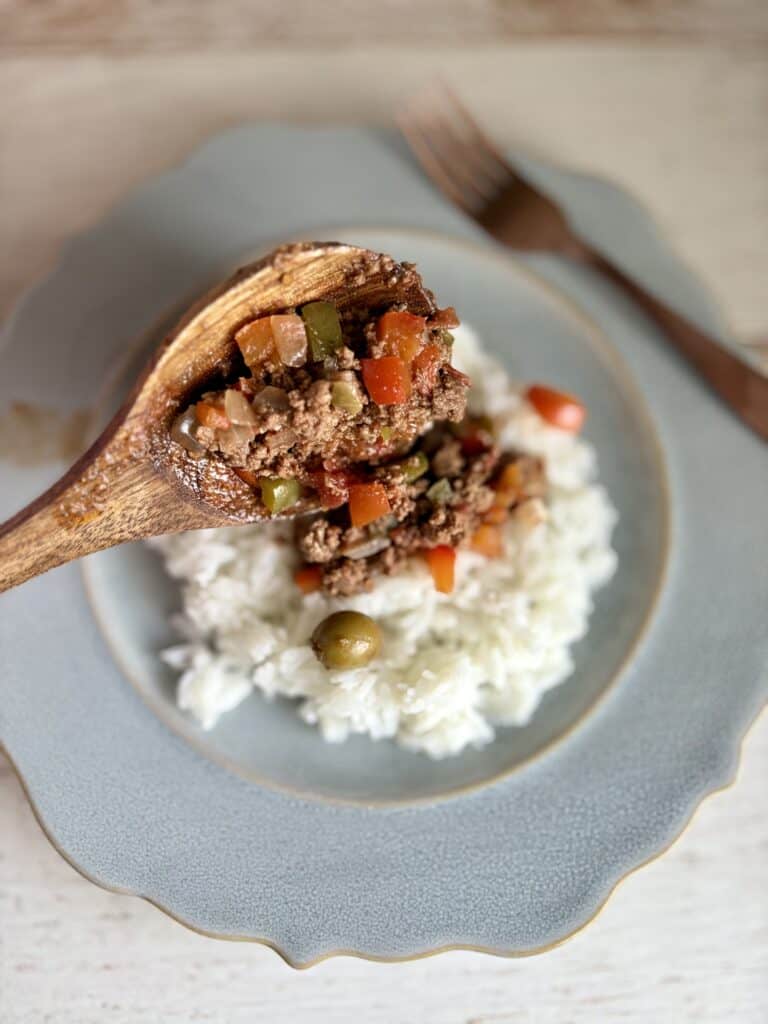 A wooden spoon holding a homemade picadillo with beef and vegetables over a bed of white rice on a blue plate.