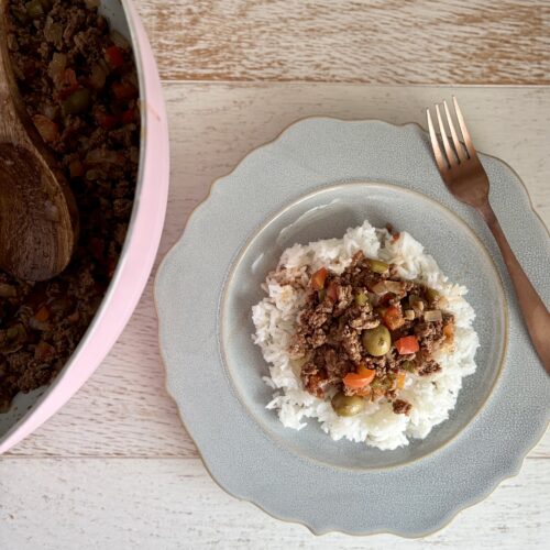 A plate of homemade beef picadillo served over rice, with a fork on the side, on a light wooden table.
