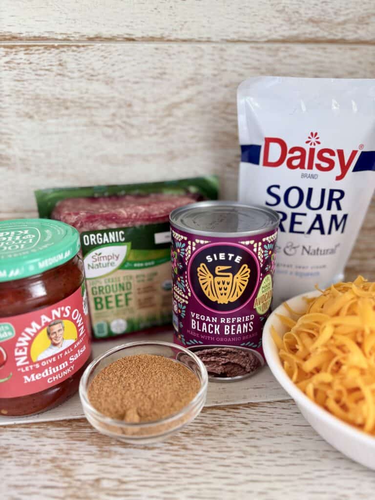 Ingredients for baked taco dip dish including black beans, sour cream, ground beef, and cheese on a wooden surface.