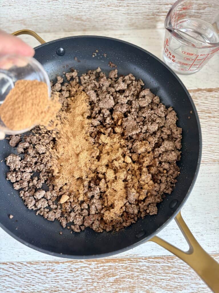 Ground beef being cooked in a skillet with spices and water nearby, ready for a delicious meal.