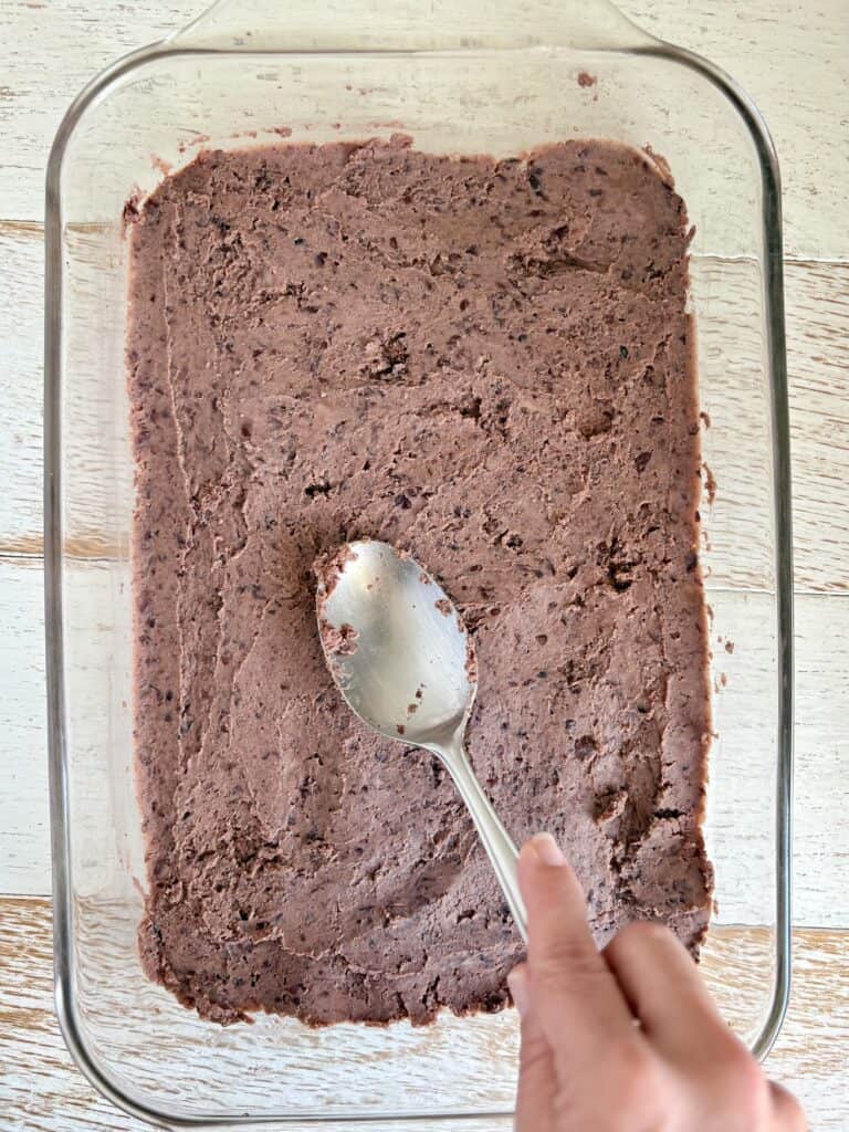 A glass baking dish filled with refried black beans being spread with a spoon, ready to set, on a rustic wooden surface.