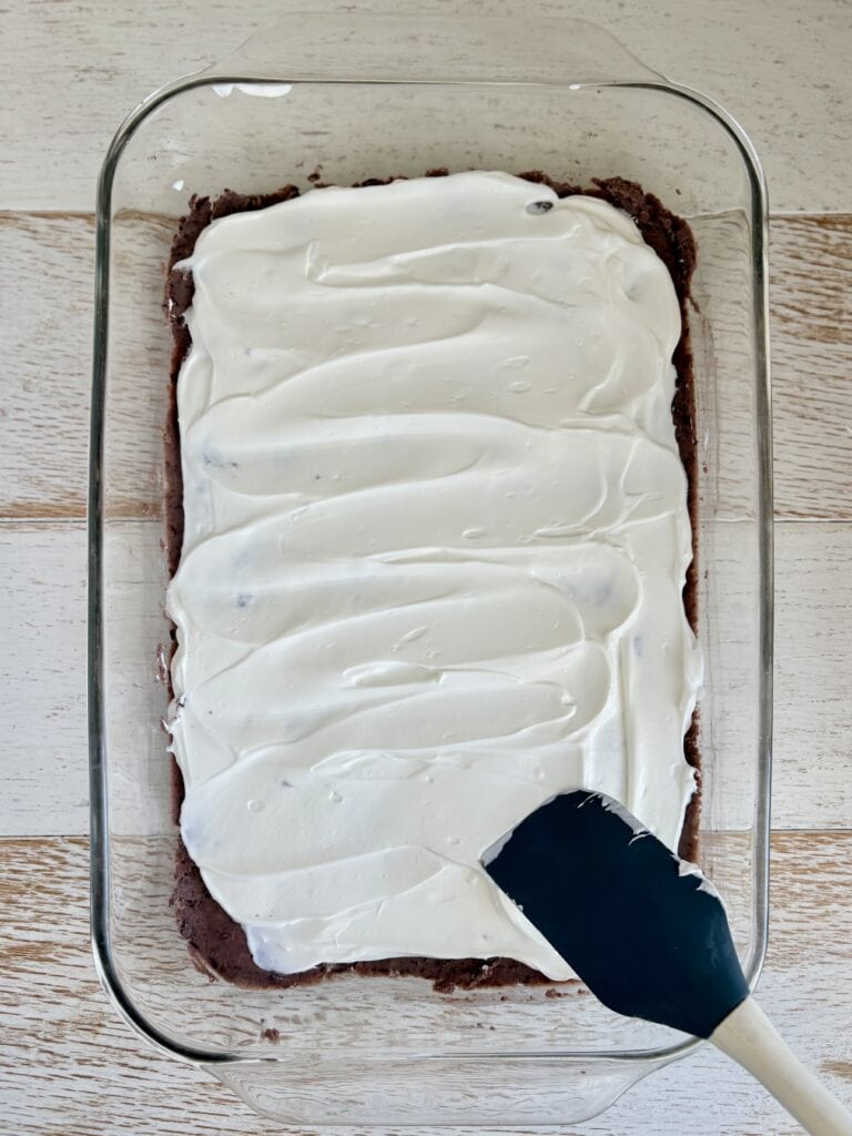 Sour cream being spread over a layer of refried black beans in a glass baking dish