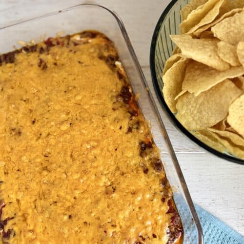 Baked Layered Taco Dip in a glass dish next to a bowl of tortilla chips on a wooden counter.