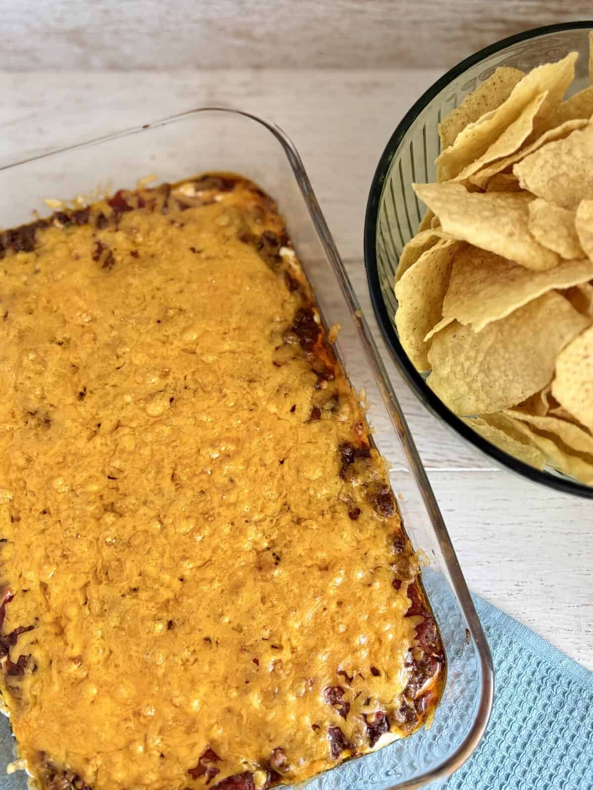 Baked Layered Taco Dip in a glass dish next to a bowl of tortilla chips on a wooden counter.