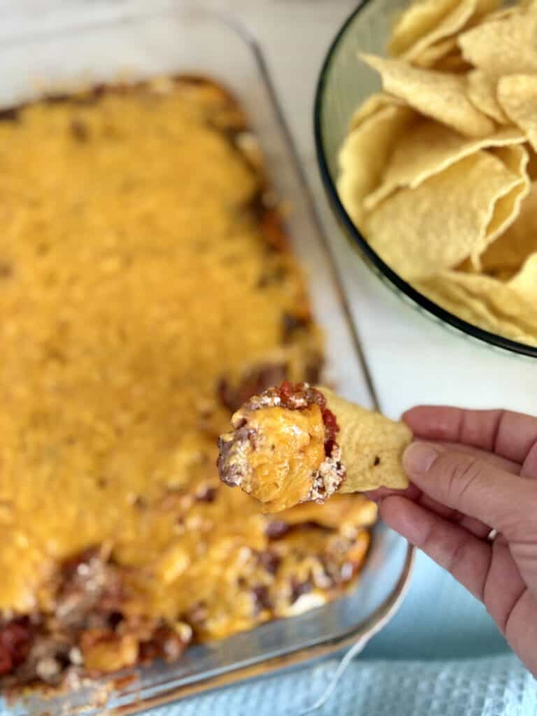 Close-up of a cheesy baked dish in a glass baking dish with a hand holding a chip topped with cheese and meat, and a bowl of tortilla chips in the background.