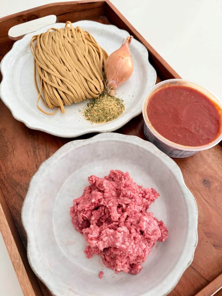 Whole wheat fresh linguine on a plate with Mrs. Dash garlic and herb seasoning and a shallot on a gray plate in front of that is tomato sauce in a plastic container and in front of that a gray plate is 8 ounces of ground beef all on a brown tray on the white counter