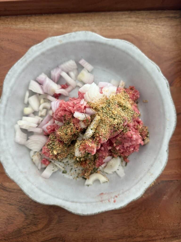 Ground meat, diced shallots, and seasoning in a grey plate on a brown tray.