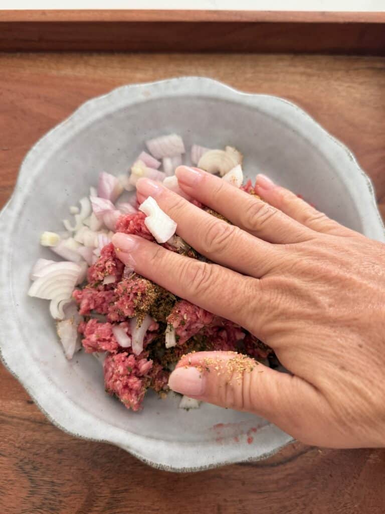 Using clean hands to mix meat, seasoning and diced shallots in a grey plate on a brown tray.
