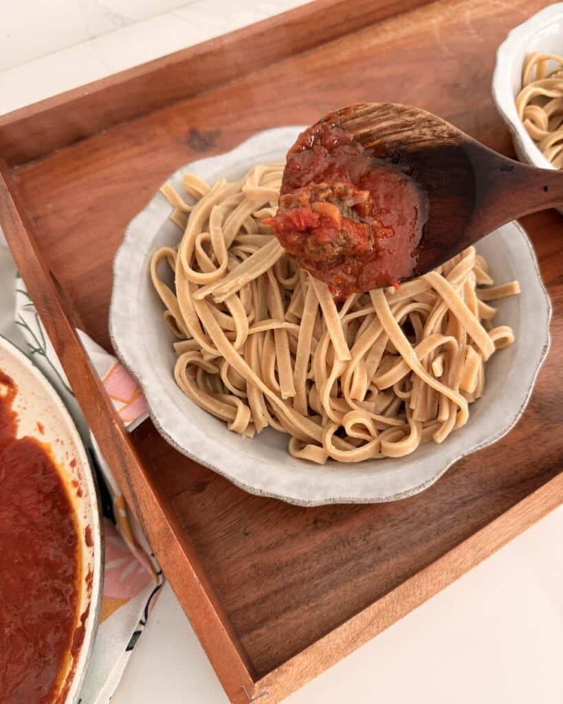 Easy Linguine and Meatballs for two in a grey plate on a brown tray with a wooden spoon pouring tomato sauce onto the linguine.