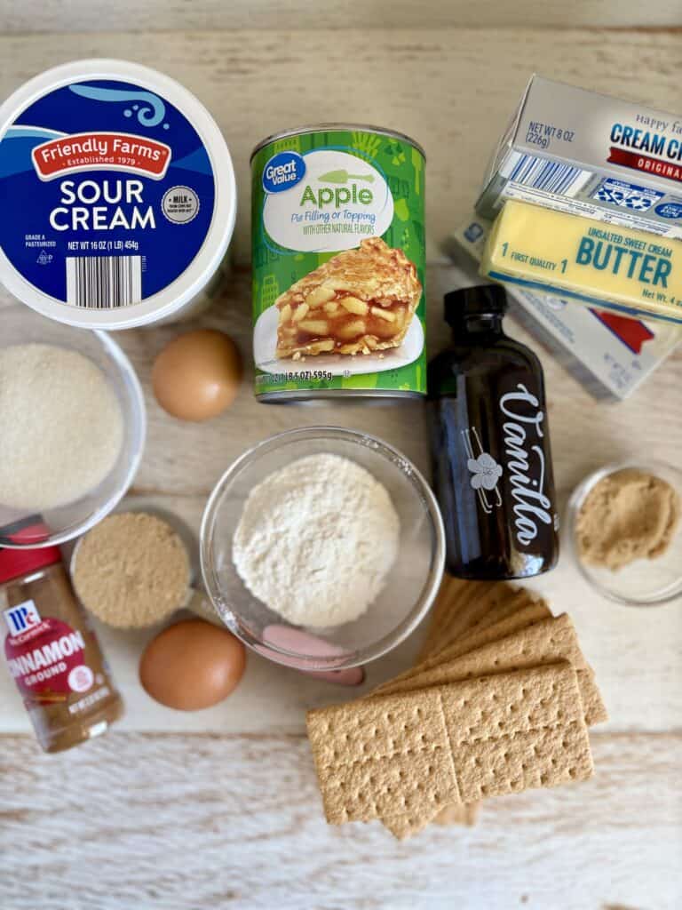 Assorted ingredients for apple cheesecake bars including sour cream, eggs, butter, apple filling, cinnamon, and graham crackers on a wooden surface.