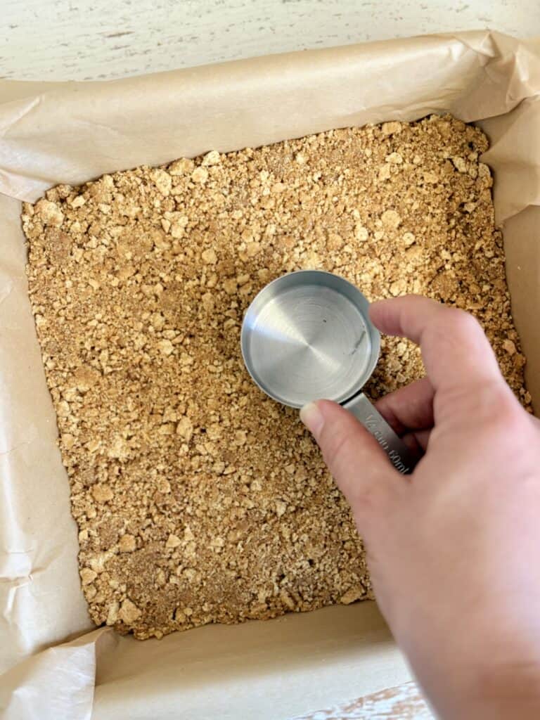 Close-up of a hand holding a metal measuring cup pressing a graham cracker crust into a square pan.
