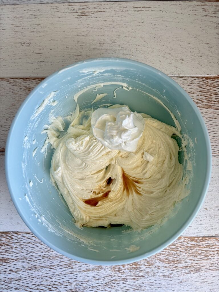 Close-up of smooth, creamy cheesecake batter in a light blue mixing bowl with a dollop of sour cream and vanilla ready for mixing