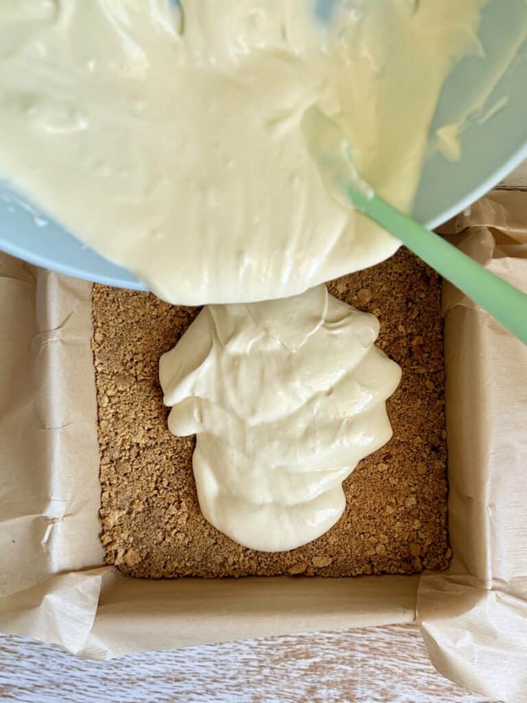 Creamy cheesecake filling being poured over a graham cracker crust in a baking pan, ready for baking or chilling.