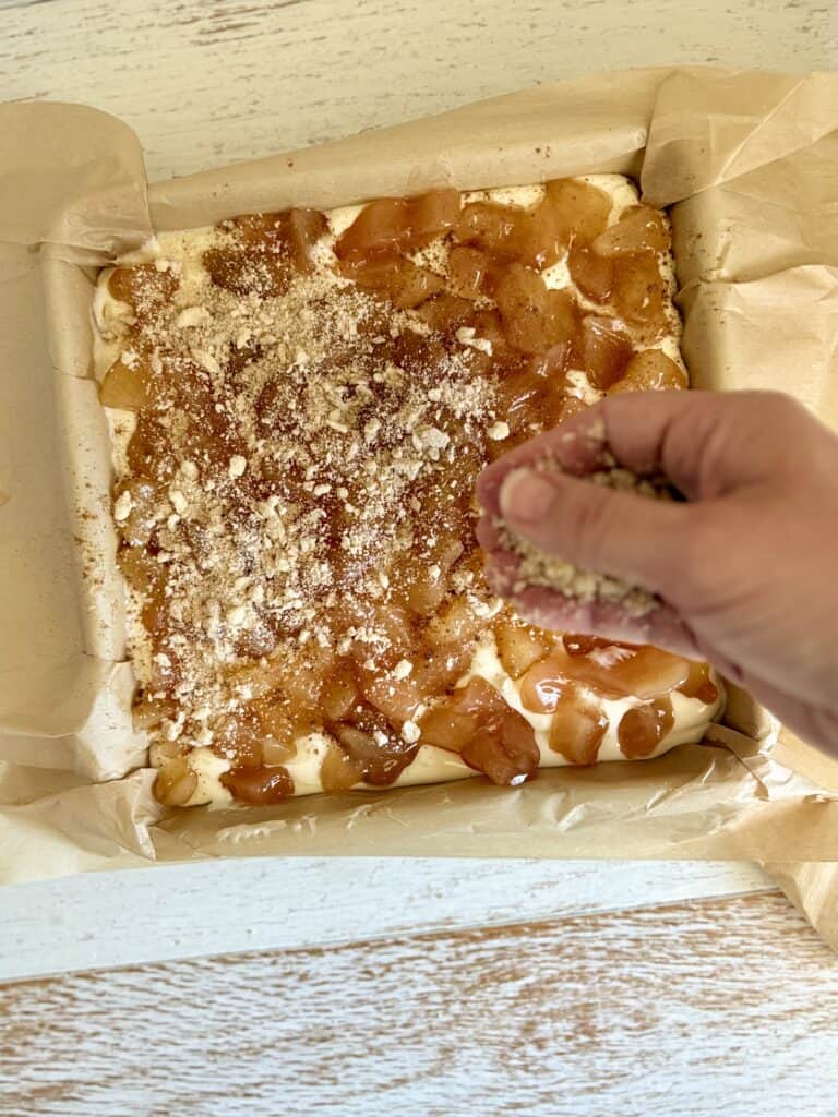 A hand sprinkling streusel over the apple layer of cheesecake in a parchment-lined baking pan.