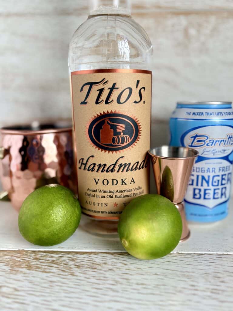 Bottle of Tito's Handmade Vodka with fresh limes and cocktail accessories on a rustic wooden surface.