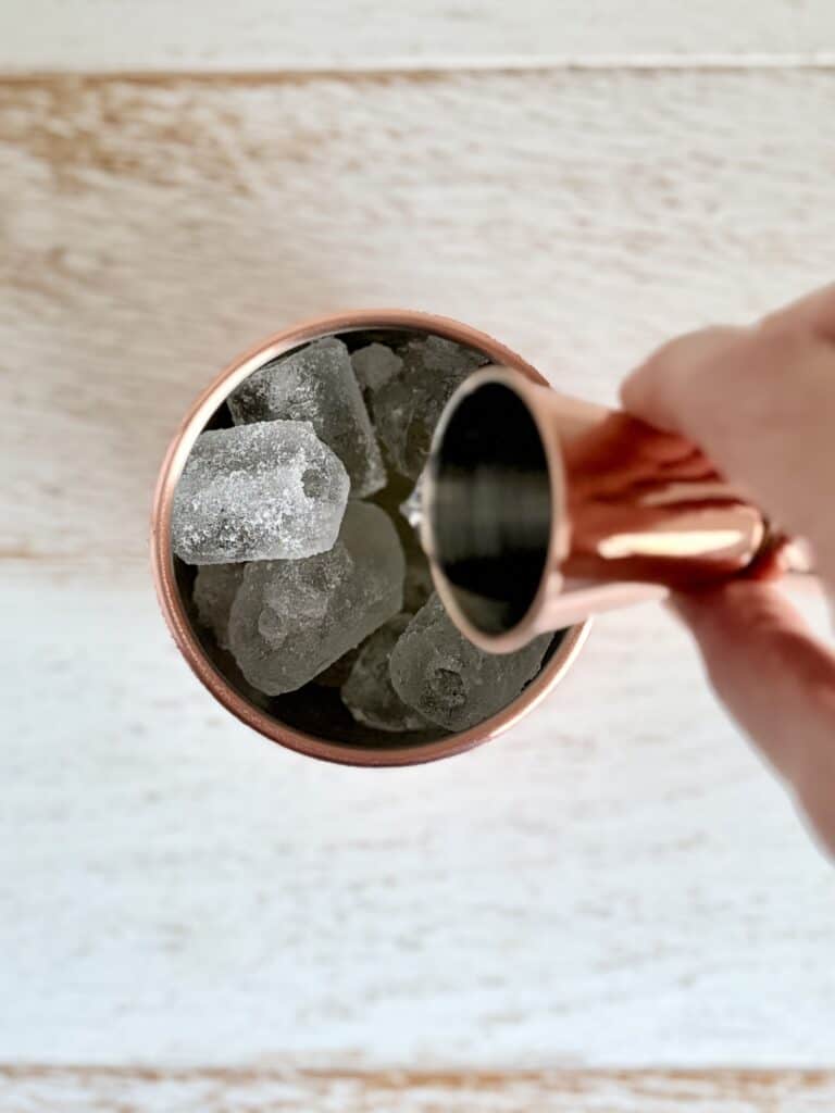 Copper Moscow Mule mug filled with ice cubes, held by a hand, on a light wooden surface, showcasing a refreshing drink presentation.