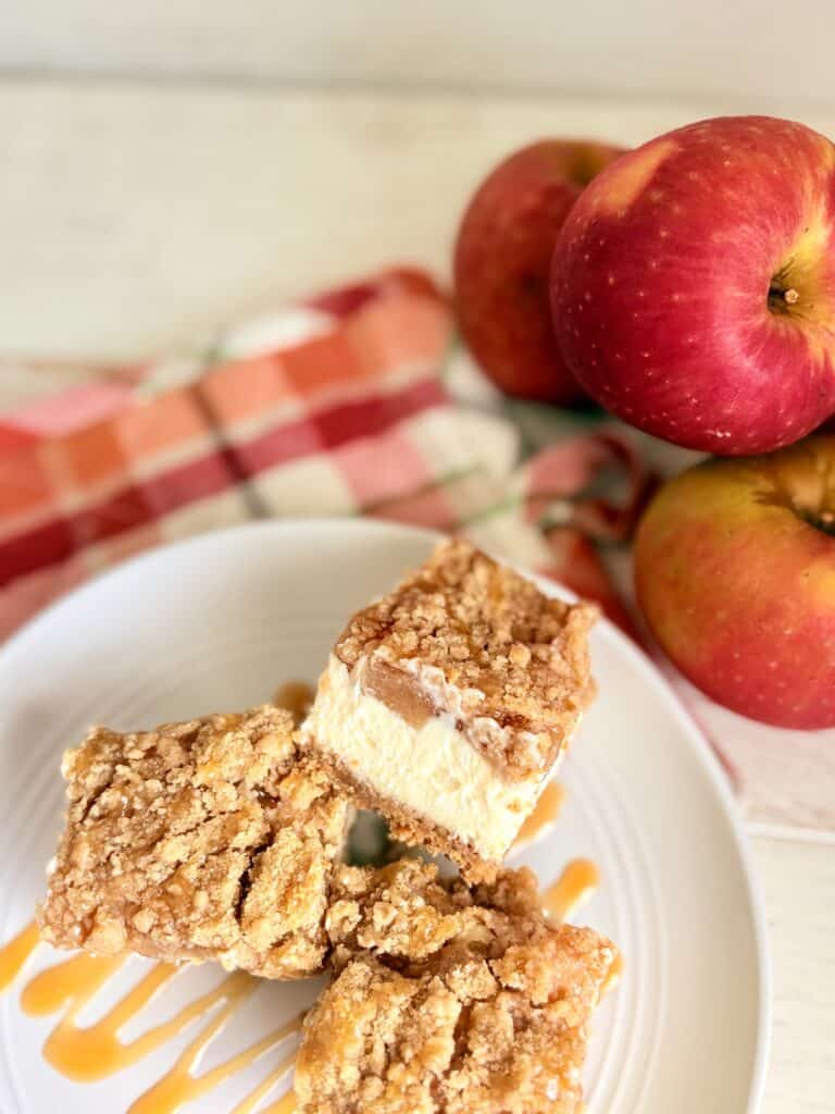 Caramel Apple Cheesecake Bars on a white plate drizzled with more caramel next to fresh apples.