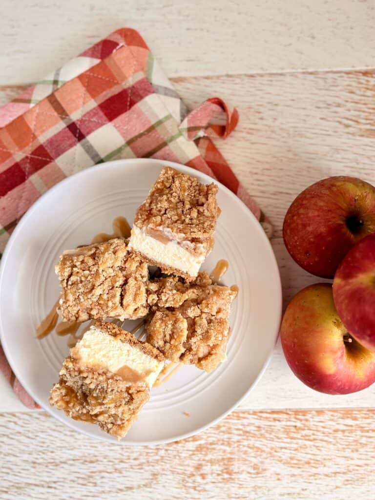 A white plate with caramel apple cheesecake bars on it next to apples and a fall themed pot holder.
