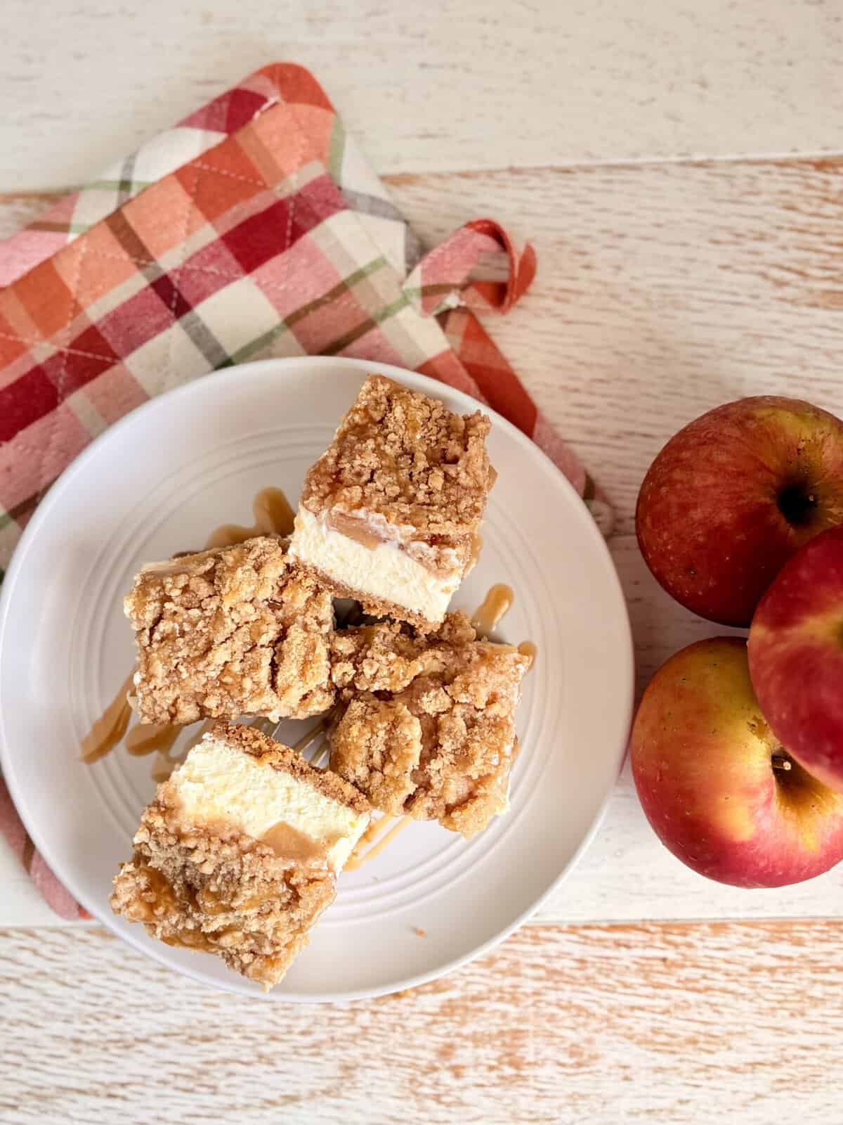 A white plate with caramel apple cheesecake bars on it next to apples and a fall themed pot holder.