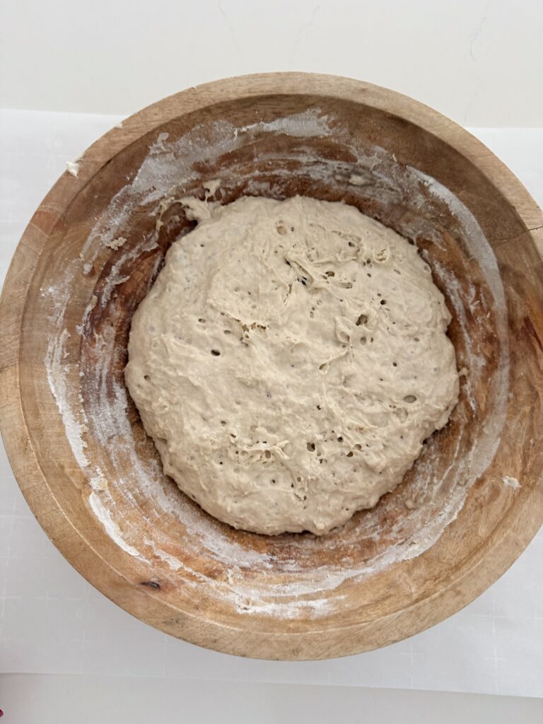 Risen no-knead bread dough with a bubbly surface in a wooden bowl.