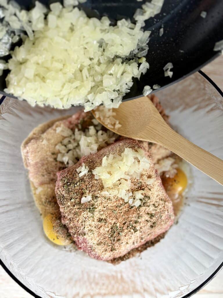 Cooked onions being scooped into a bowl with a meat, egg and breadcrumb mixture.