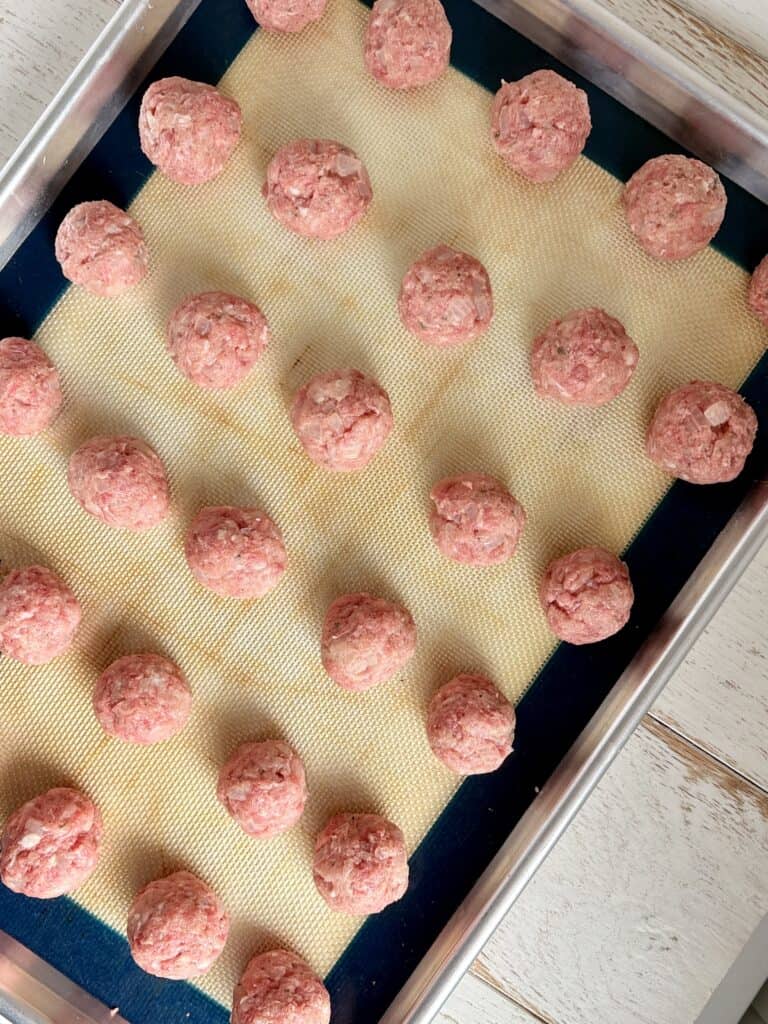 A baking sheet lined with a silicone mat holding rolled meatballs ready to be baked.