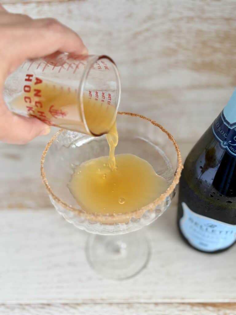 Apple cider being poured into a coupe glass with a cinnamon sugar rim next to a bottle of Prosecco