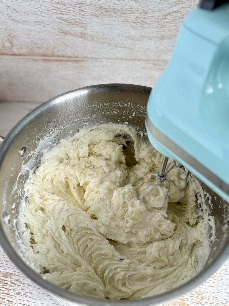 Creamy mashed potatoes being prepared in a stainless steel mixer bowl with a hand mixer attached, on a wooden surface.