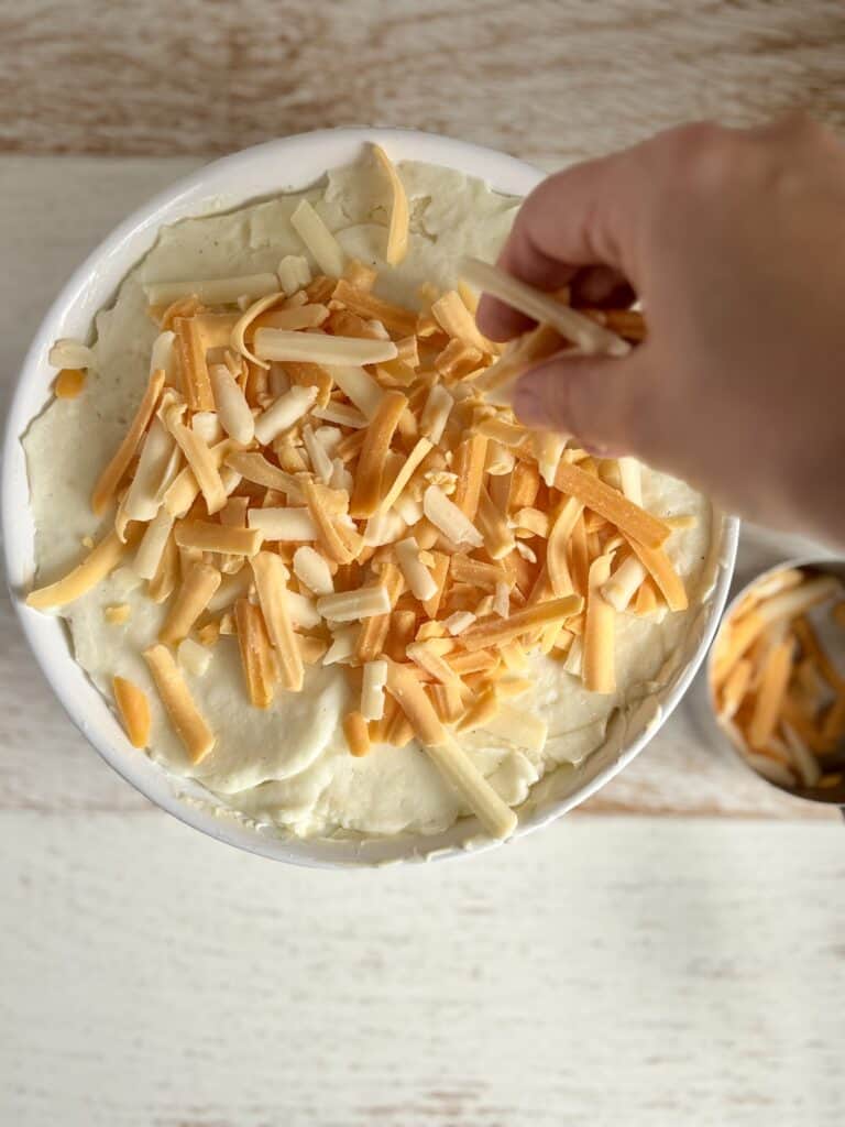 Person sprinkling shredded cheese over creamy mashed potatoes in a white bowl, ready to be baked or served.