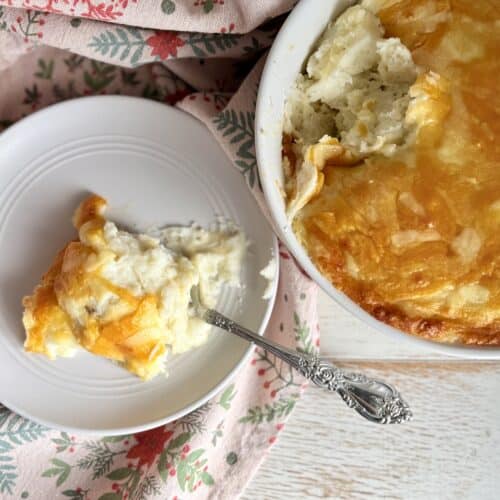 Cheesy mashed potatoes in a bowl next to a white plate with a silver spoon.