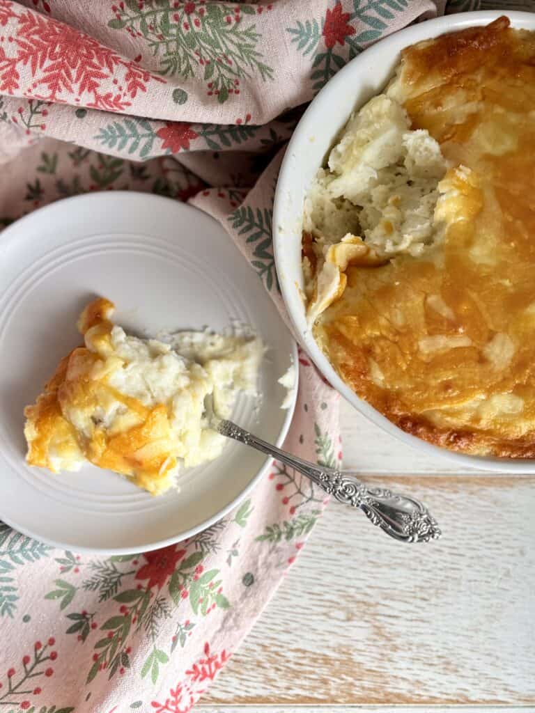 Cheesy mashed potatoes in a bowl next to a white plate with a silver spoon.