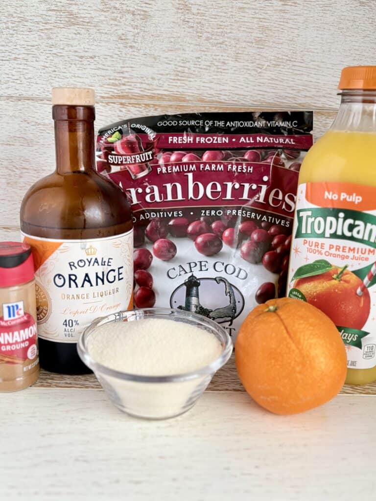 Assorted fresh fruits, orange liquor, and juice on a light wooden surface, including strawberries, an orange, and a glass of cream for cooking or smoothies.