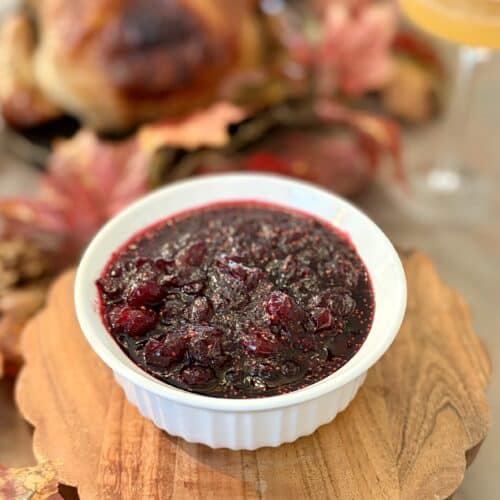 Close-up of homemade cranberry sauce in a white bowl, served on a wooden platter with a roasted turkey and autumn leaves in the background.