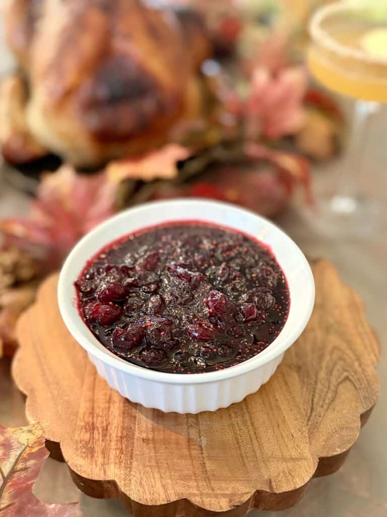 Close-up of homemade cranberry sauce in a white bowl, served on a wooden platter with a roasted turkey and autumn leaves in the background.