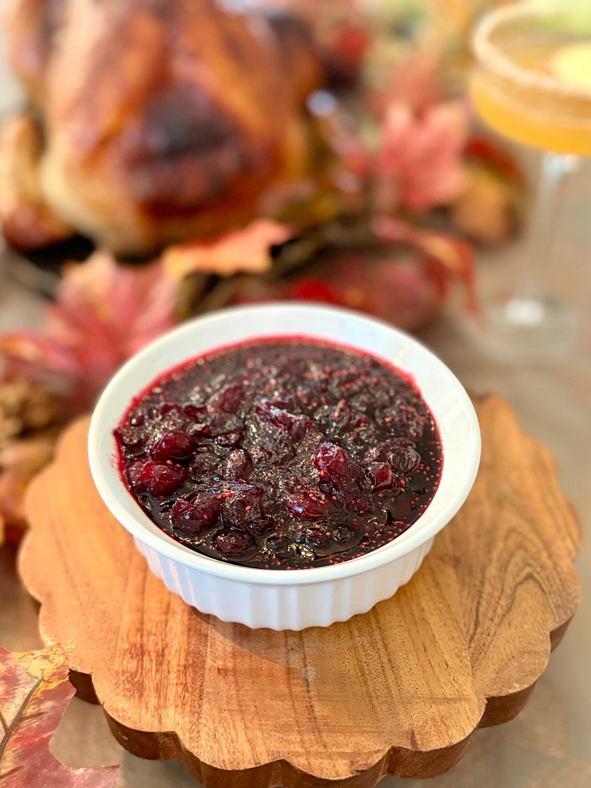 Close-up of homemade cranberry sauce in a white bowl, served on a wooden platter with a roasted turkey and autumn leaves in the background.
