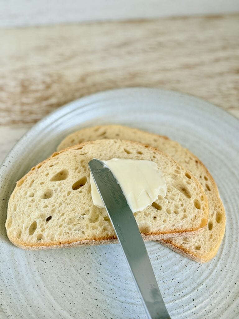 Slice of rustic bread with a pat of butter and a butter knife on a light ceramic plate, perfect for breakfast or snack.